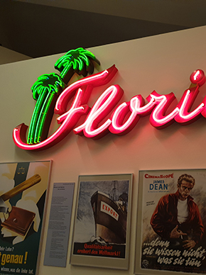 Teil einer Leuchtreklame mit dem roten Schriftzug Florida und zwei grünen Palmen, die an einer Wand in der Dauerausstellung über verschiedenen Postern hängt., auf denen unter anderem James Dean zu sehen ist.