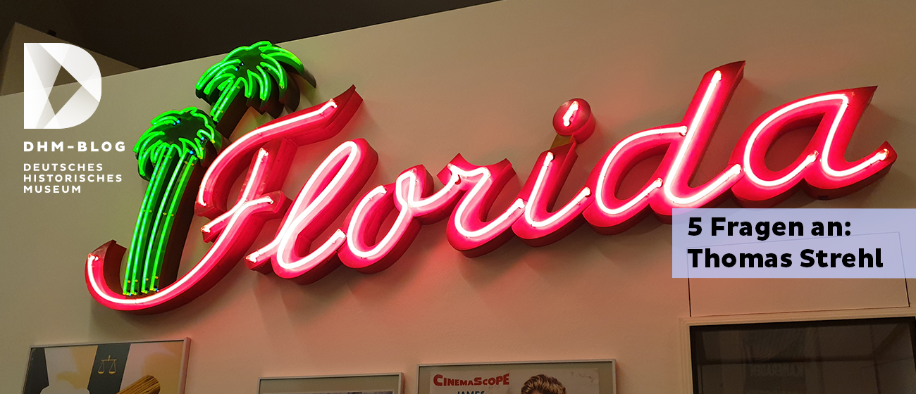 Eine Leuchtreklame mit dem roten Schriftzug Florida und zwei grünen Palmen, die an einer Wand in der Dauerausstellung über verschiedenen Postern hängt.