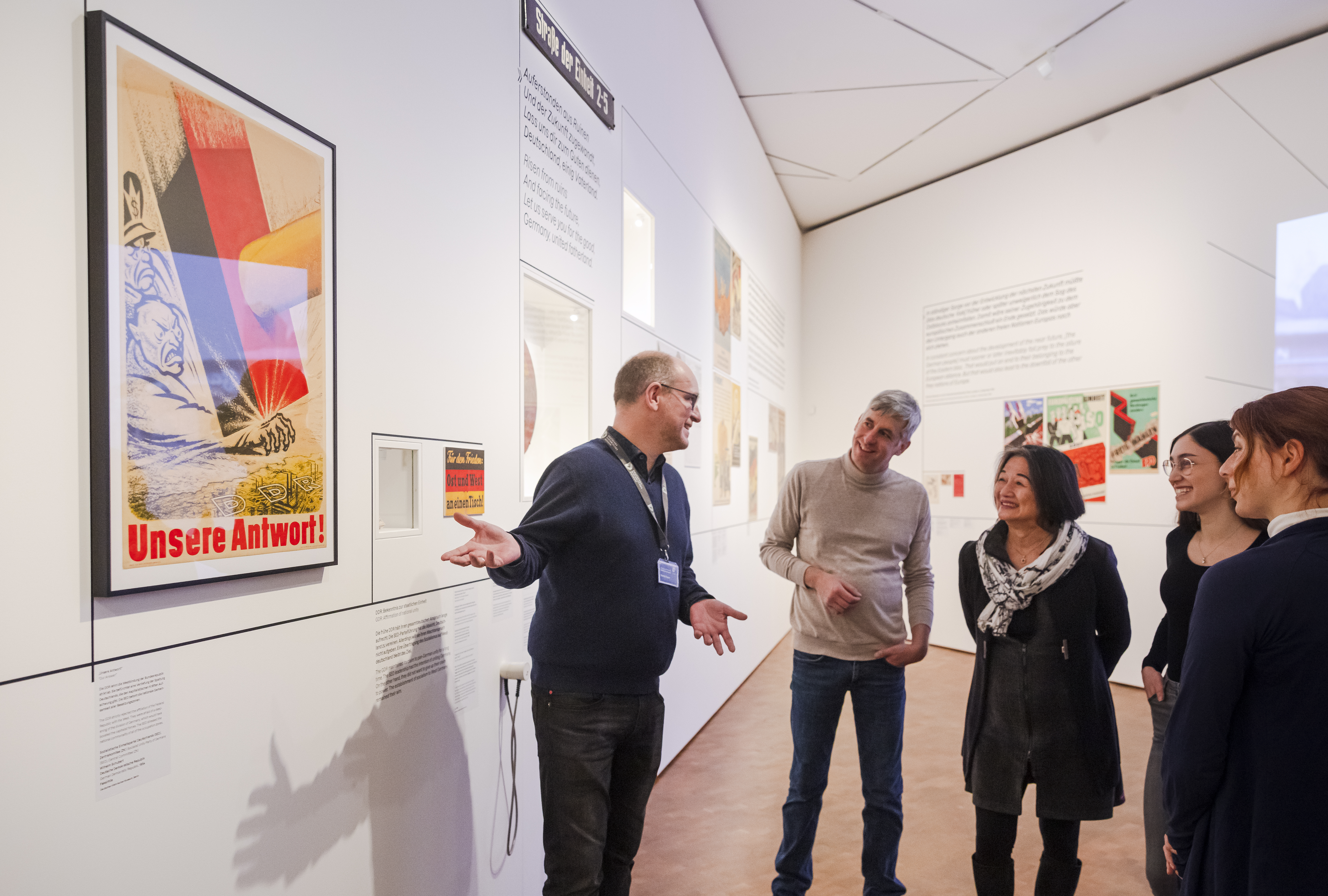 Ein Mann steht vor einer Ausstellungswand und spricht zu einer Gruppe.