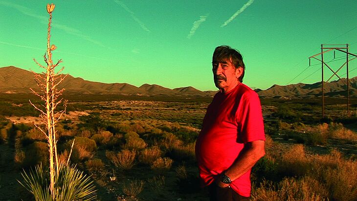 Ein Mann mit rotem T-Shirt steht in einer wüstenähnlichen Landschaft und blickt in die Kamera.