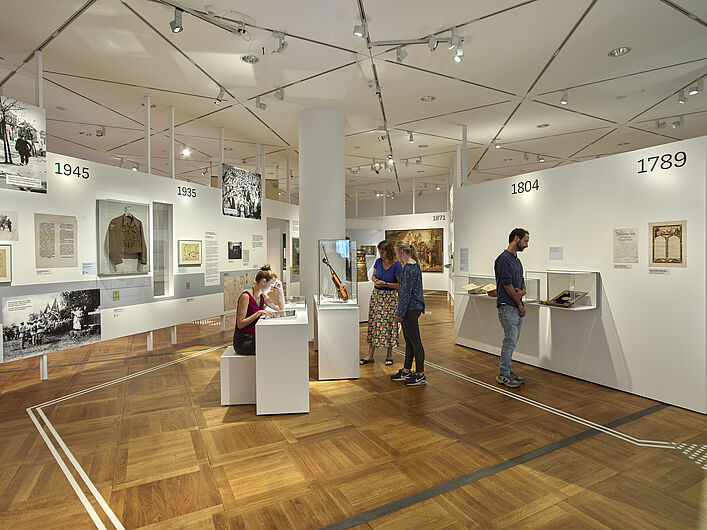 Das Bild zeigt einen Einblick in die Ausstellung. Es sind mehrere Personen zu sehen, die sich unterschiedliche Objekte ansehen. Zwei Personen gucken auf eine Violine, die in einer Vitrine aufbewahrt wird. Eine Person sitzt an einem Tisch und liest etwas, eine andere Personen steht an der Wand und guckt sich einen Text an.