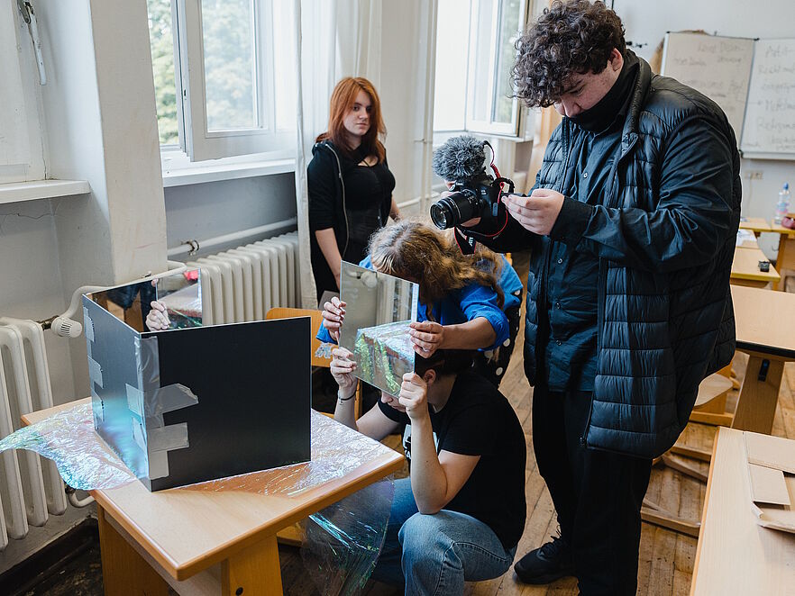 Schüler filmt in einem Klassenzimmer mit einer Kamera, wie eine Mitschülerin verspiegelte Platten über ein selbstgebautes Objekt auf einem Schultisch hält. Im Hintergrund steht eine Mitschülerin mit roten Haaren am offenen Fenster.
