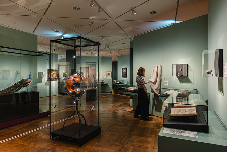 Ausstellungsansicht "Natur und deutsche Geschichte. Glaube – Biologie – Macht" Eine Frau in gestreiftem Oberteil fühlt an einem weißen Baumwolltuch