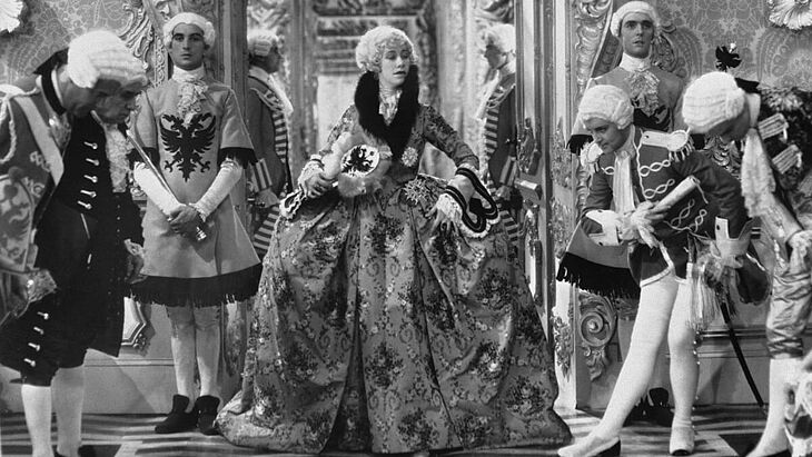 Eine Frau in einem historischen Gewand und Männer, die um sie herum stehen und sich verbeugen.