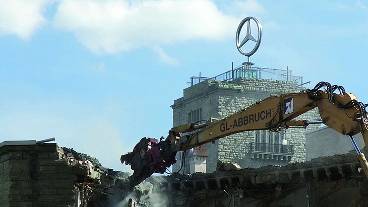 Eine Baustelle mit Bagger vor einem Haus mit dem Mercedesstern auf dem Dach.