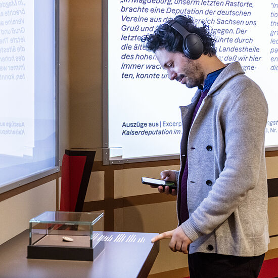 Ein Mann benutzt den Audioguide mit Kopfhörern und Handy und betrachtet ein Objekt auf einem Tisch.