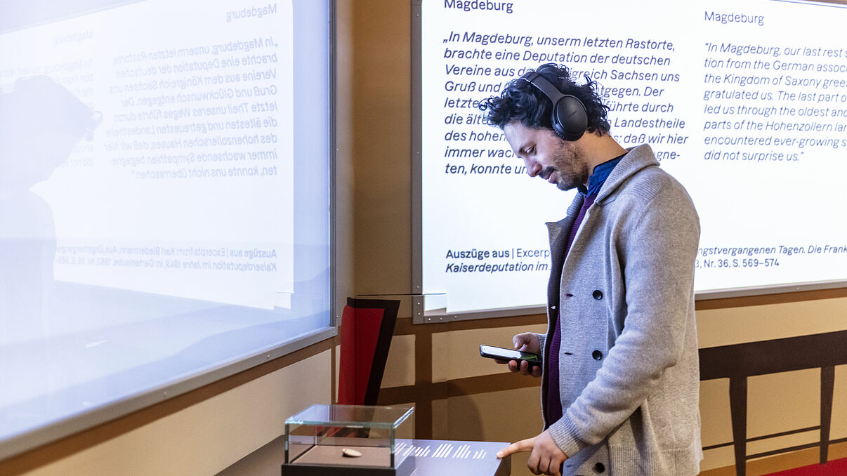 Ein Mann benutzt den Audioguide mit Kopfhörern und Handy und betrachtet ein Objekt auf einem Tisch.