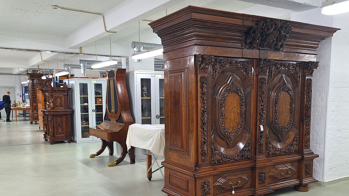 Ein alter Schrank steht in einem Depot und weitere alte Möbel sind zu sehen
