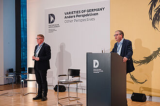 Zwei Männer in Anzügen auf einer Bühne mit Podium und Stühlen, im Hintergrund Schriftzug 