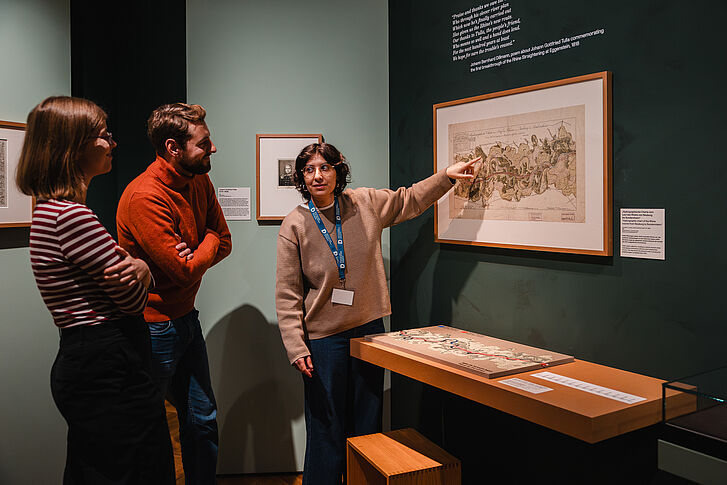 Ausstellungsansicht "Natur und deutsche Geschichte. Glaube – Biologie – Macht" Eine Frau in einem beigen Pullover gibt zwei Besuchenden eine Führung und zeigt auf ein Bild
