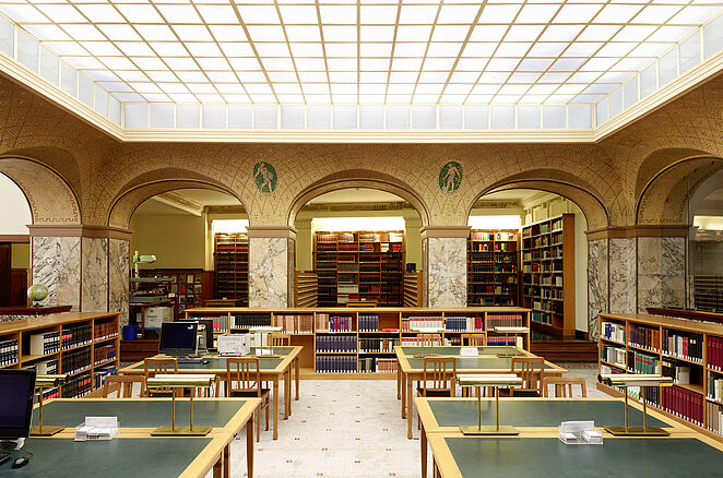 Auf dem Bild ist das Innere der Bibliothek zu sehen. Die Tische sind grün und hintereinander angerichtet. Im Hintergrund sind viele Bücher in Regalen zu sehen.