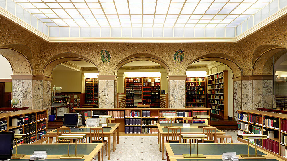 Auf dem Bild ist das Innere der Bibliothek zu sehen. Die Tische sind grün und hintereinander angerichtet. Im Hintergrund sind viele Bücher in Regalen zu sehen.