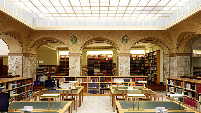 Auf dem Bild ist das Innere der Bibliothek zu sehen. Die Tische sind grün und hintereinander angerichtet. Im Hintergrund sind viele Bücher in Regalen zu sehen.