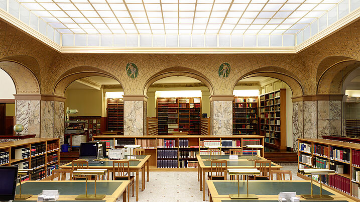 Auf dem Bild ist das Innere der Bibliothek zu sehen. Die Tische sind grün und hintereinander angerichtet. Im Hintergrund sind viele Bücher in Regalen zu sehen.
