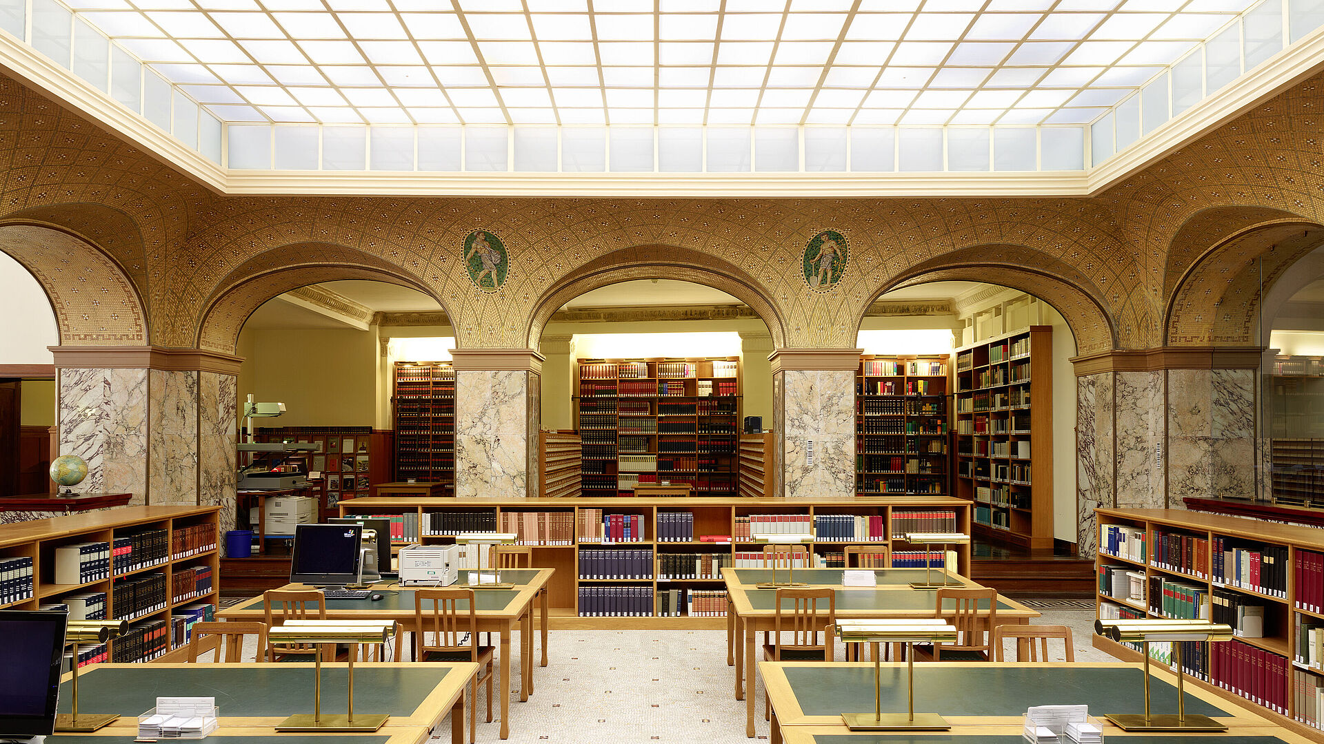 Auf dem Bild ist das Innere der Bibliothek zu sehen. Die Tische sind grün und hintereinander angerichtet. Im Hintergrund sind viele Bücher in Regalen zu sehen.