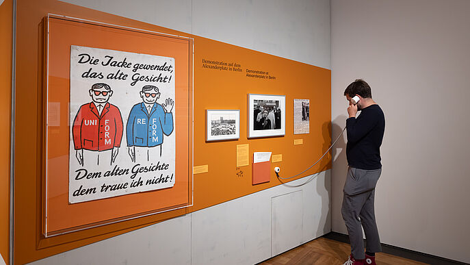 Ein Mann hört sich ein Audio an und sieht sich dabei etwas an einer Ausstellungswand an.