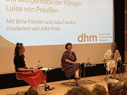 Drei Personen sitzen auf einer Bühne und sprechen miteinander vor Publikum.