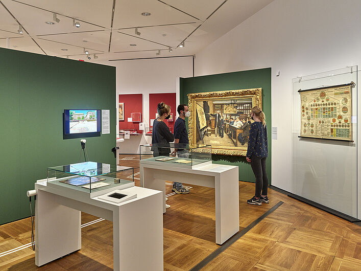 Auf dem Bild ist eine Szene der Ausstellung zu sehen. Im Hintergrund stehen Personen um ein Objekt herum. Im Vordergrund sind zwei Vitrinen zu sehen, in denen sich Objekte befinden.