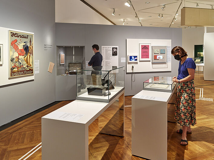 Auf dem Bild ist eine Szene in der Ausstellung zu sehen. Die Wände sind grau. Auf den grauen Wändens idn verschiedene Objekte zu sehen. Zum Beispiel ein großes Plakat mit einem Hahn drauf. Im Vordergrund sieht sich eine Person eine Vitrine an, in der ein Dokument gezeigt wird.