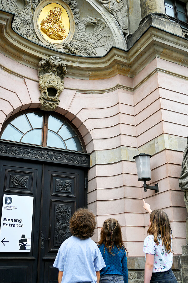 Von Drachen und Kronen. Mit dem Fernglas rund ums Zeughaus Foto von drei Kindern vor dem Zeughaus. Ein Mädchen zeigt mit dem Finder auf ein Relief.