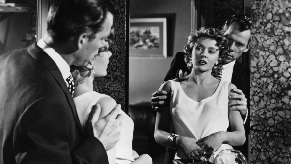 Ein Mann steht hinter einer Frau und greift ihre Arme, während die beiden sich im Spiegel betrachten.