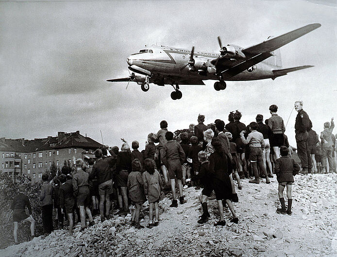 Henry Ries, Landeanflug eines „Rosinenbombers“ in Tempelhof, Juli 1948. Eine viermotorige Douglas-Maschine C-54 Skymaster, der sogenannte Rosinenbomber, im Landeanflug auf den Flughafen Tempelhof. Die C-54 war das Haupttransportflugzeug der Luftbrücke. Auf einem Schuttberg haben sich viele Berliner Kinder und Jugendliche versammelt, um die Flugzeuge starten und landen zu sehen. (Inv.Nr. Ph 2008/730)