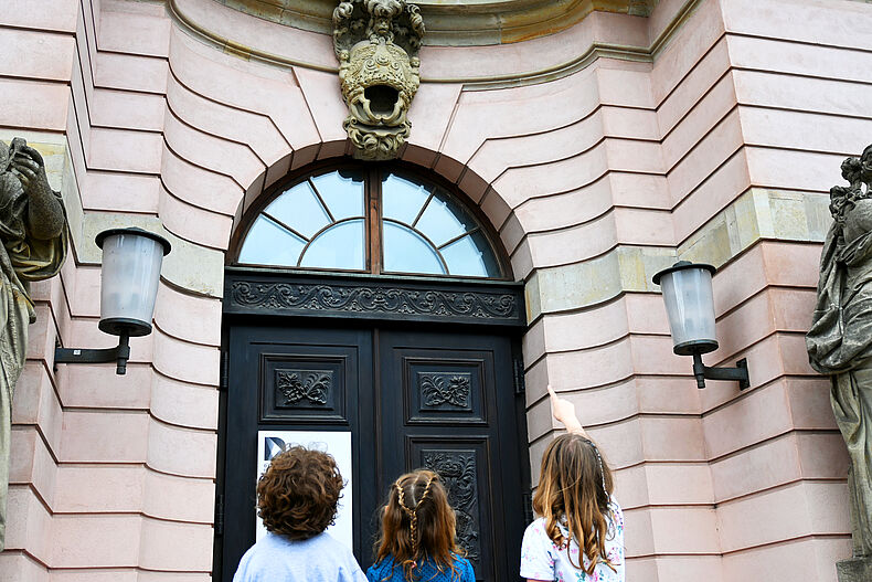 Von Drachen und Kronen. Mit dem Fernglas rund ums Zeughaus Foto von drei Kindern vor dem Zeughaus. Ein Mädchen zeigt mit dem Finder auf ein Relief.