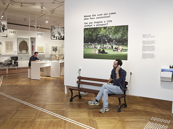 Auf dem Bild ist ein Teil der Ausstellung "Staatsbürgerschaften" zu sehen. Vor einer Wand mit eienm Parkfoto sitzt ein Mann. Er hat eine Hörmuschel in der Hand, die mit der Wand verbunden ist. Auf einem Text über dem Bild steht "Können Sie sich ein Le