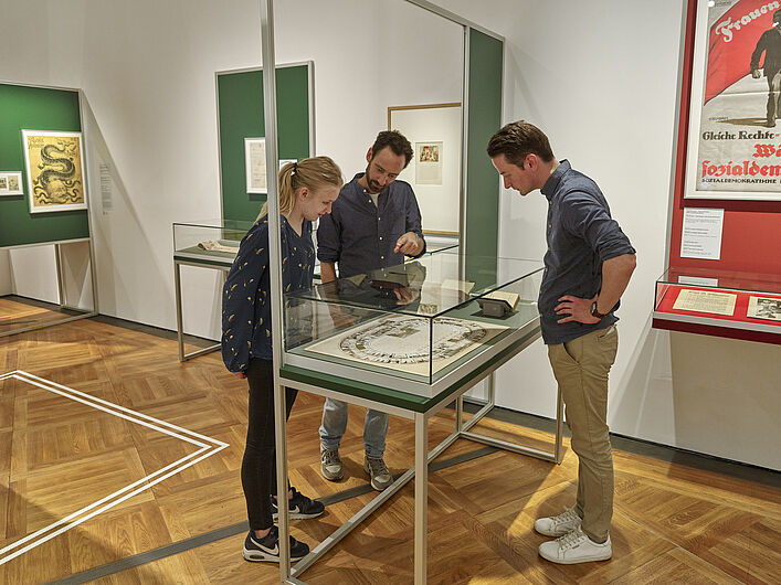 Auf dem Bild sind drei Personen zu sehen. Alle drei gucken sich ein Objekt an, das vor ihnen in einer Vitrine zu sehen ist. Einer der drei Personen zeigt auf das Objekt. Im Hintergrund hängen Bilder an der Wand.