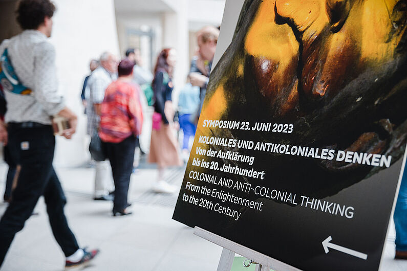 Plakat vom Symposium und stehende Menschen im Hintergrund.