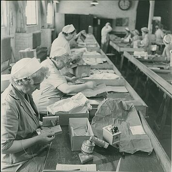 Arbeiterinnen in Uniform und mit Kopfbedeckung kontrollieren in einer Halle Gegenstände an einem langen Tisch.