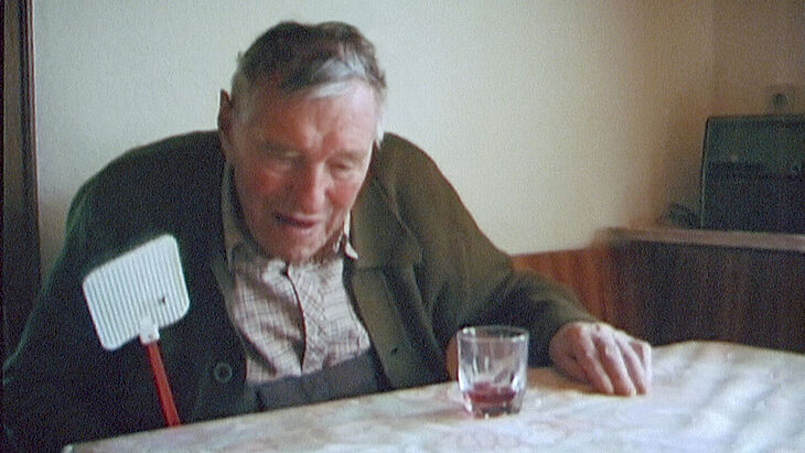 Ein älterer Herr sitzt mit einer Fliegenklatsche in der Hand an einem Tisch.