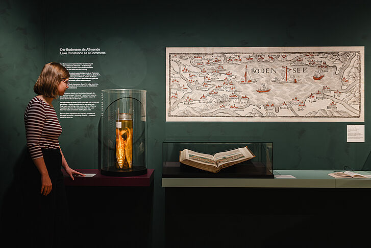 Ausstellungsansicht "Natur und deutsche Geschichte. Glaube – Biologie – Macht" Eine Frau in einem gestreiften Oberteil betrachtet Fische im Glas