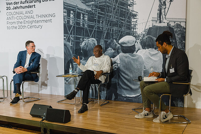 David Simo und George Steinmetz sitzen mit dem Moderator Gwinyai Machona auf der Bühne und reden.