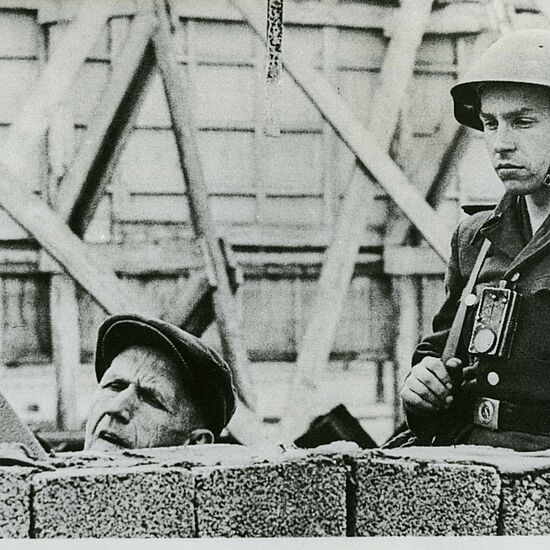 Ein Soldat in Uniform mit Helm steht neben einem Bauarbeiter, der hinter einer unvollständigen Mauer arbeitet.