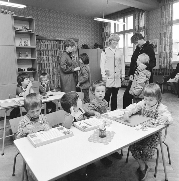 Kinder sitzen an Tischen und Frauen stehen darum und reden.