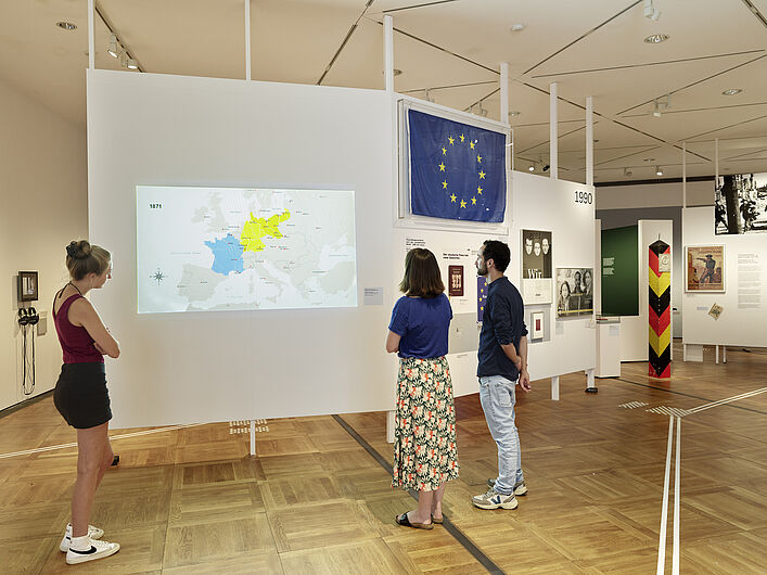 Das Bild zeigt ein Stück der Ausstellung. Drei Personen stehen vor einer Wand, auf der eine Karte abgebildet ist. Die Karte zeigt unterschiedliche Zeiten in Europa und die Entwicklung von Grenzen innerhalb Europas. Dahinter ist eien europäische Flagge zu sehen - blau mit gelben Sternen im Kreis.