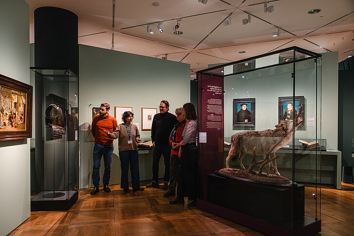 Ausstellungsansicht "Natur und deutsche Geschichte. Glaube – Biologie – Macht" Mann in rotem Pullover gibt eine Führung an vier Besuchende