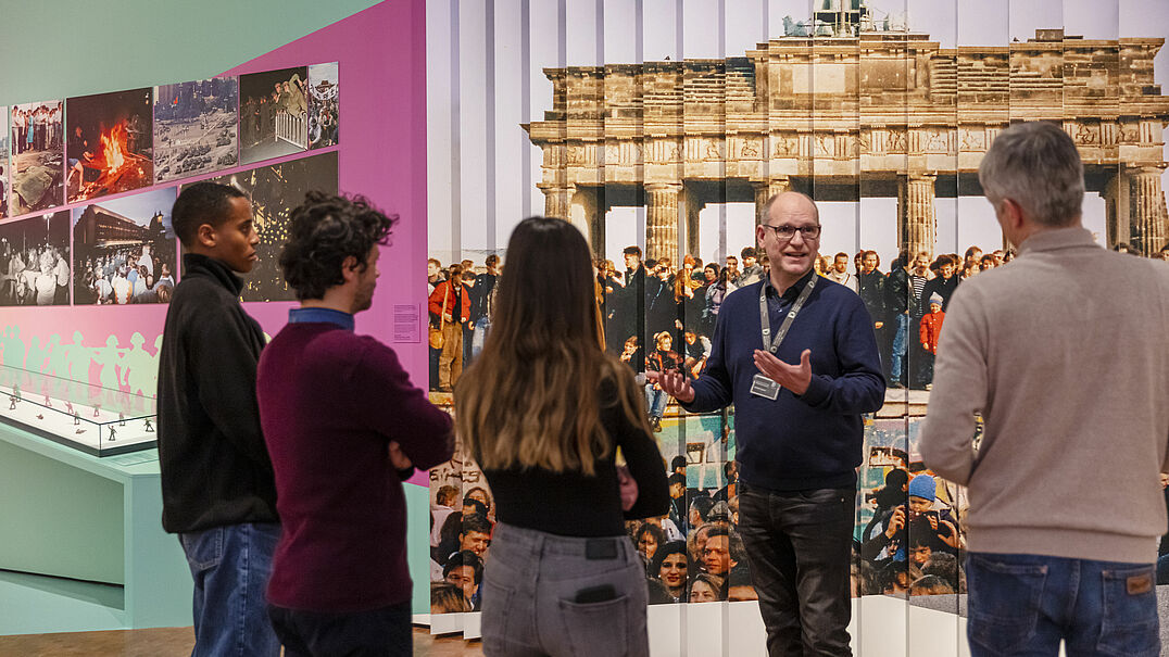 Ein Mann steht vor einer Ausstellungswand und spricht zu einer Gruppe.