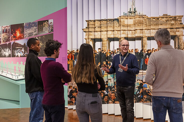 Ein Mann steht vor einer Ausstellungswand und spricht zu einer Gruppe.