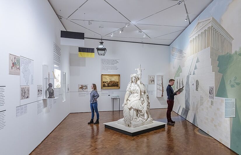 Menschen betrachten Kunstwerke in einem Museum, während sie vor einem großen Wandbild stehen.