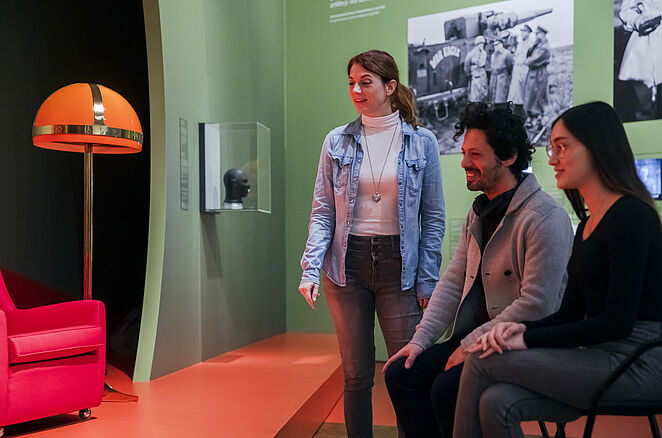 Drei Leute, von denen zwei sitzen, betrachten interessiert die Ausstellung.