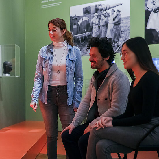 Drei Leute, von denen zwei sitzen, betrachten interessiert die Ausstellung.