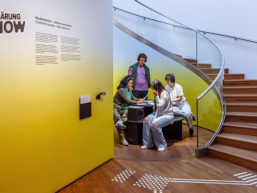 Vier Personen mit Kopfhörern in einem Museumsbereich vor einer gelbgrün leuchtenden Wand. Eine Erwachsene sitzt links an einem dunklen Tisch und hält ein Informationsblatt, ein junger Mann in lila T-Shirt steht dahinter. Rechts sitzen zwei Jugendliche am Tisch. Im Hintergrund führt eine geschwungene Glastreppe mit Holzstufen nach oben; auf den Stufen sind mehrsprachige Fragen angebracht, etwa „Wie viel Erziehung braucht…“ und „What is…“.
