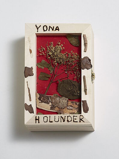 Holzschaukasten mit rotem Deckel auf weißem Hintergrund, von oben fotografiert. Oben in den hellen Holzrahmen eingebrannt der Name „YONA“, unten die Aufschrift „HOLUNDER“. Der Rahmen ist außen mit unregelmäßigen Rindenstücken und kleinen Zweigen dekoriert.