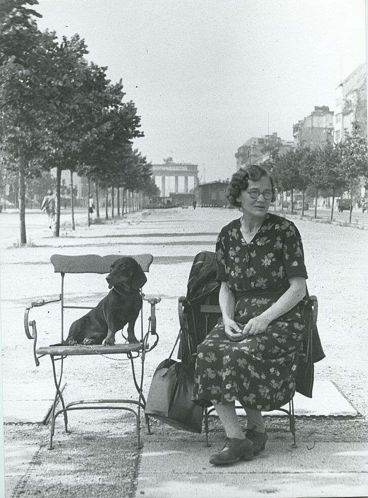 Eine Frau und ein Hund sitzen auf einer Straße mit Brandenburger Tor im Hintergrund auf Stühlen.