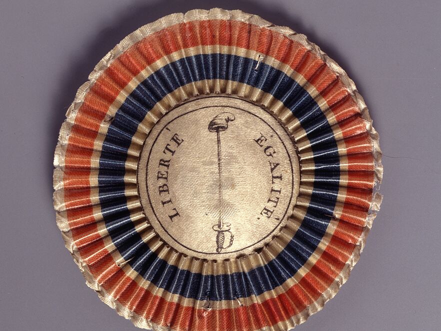 Gefalteter Stoff in Rot, Blau und Weiß; Mittelmedaillon mit Freiheitslanze und Mütze (Pike und Jakobinermütze) und Schriftzug „LIBERTÉ ÉGALITÉ.“