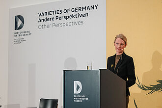 Frau in schwarzem Blazer steht hinter einem Rednerpult mit Logo des Deutschen Historischen Museums, im Hintergrund Wand mit Schrift 