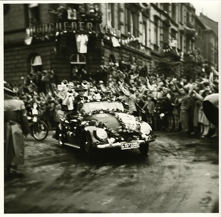 Menschenmassen jubeln ein mit Blumen geschmücktes Auto an.