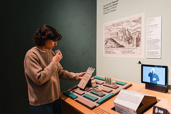 Exhibition view "Nature and German History. Faith – Biology – Power" A woman in a beige sweater smells a container.
